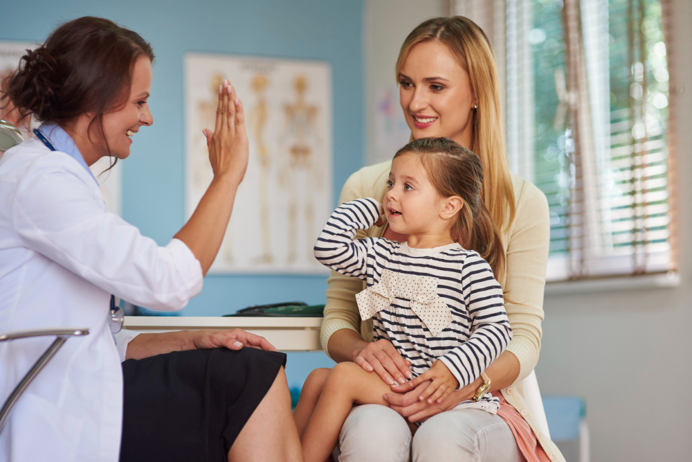 A smiling doctor gives a high five to a little girl sitting on her mother’s lap