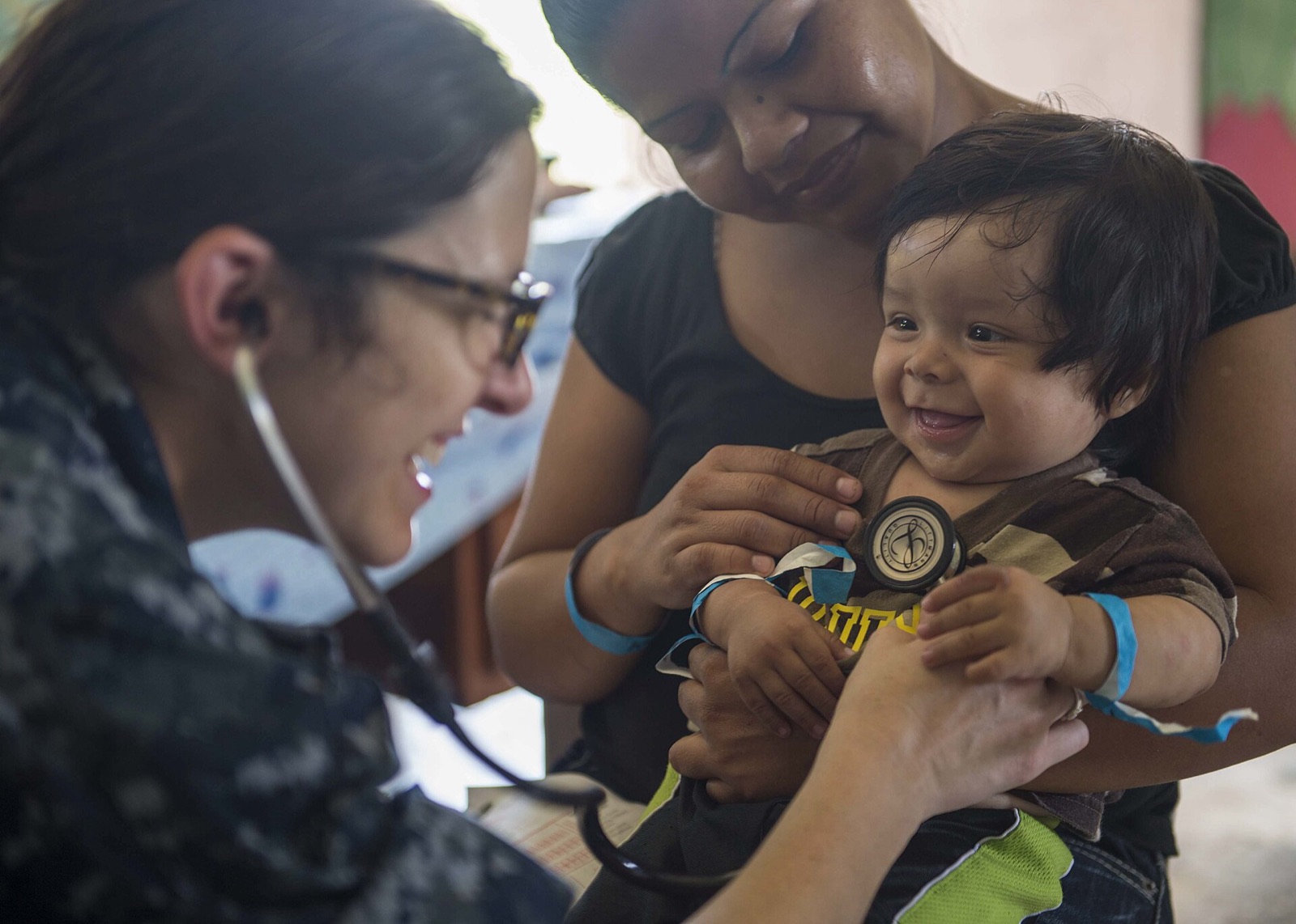 A provider listens to a smiling baby’s heartbeat while the mother holds him